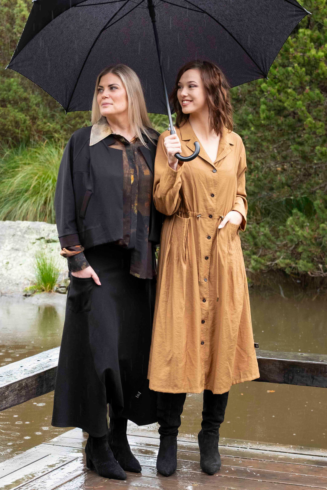 Two women standing under a black umbrella by a pond with greenery in the background wearing fashion from Deeanne Hobbs