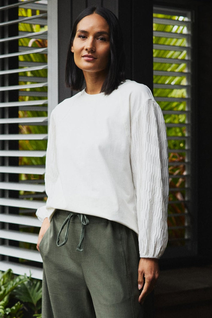 Woman wearing a white top and green pants standing in front of a window with plants outside.