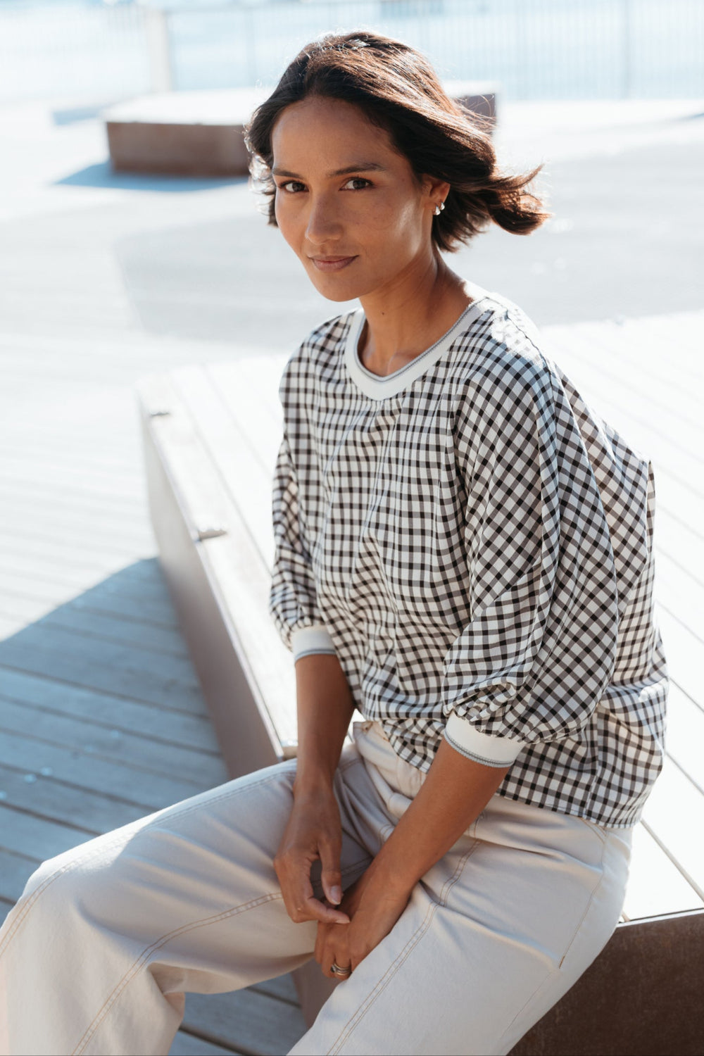Woman sitting on a wooden deck wearing a checkered shirt and white pants.