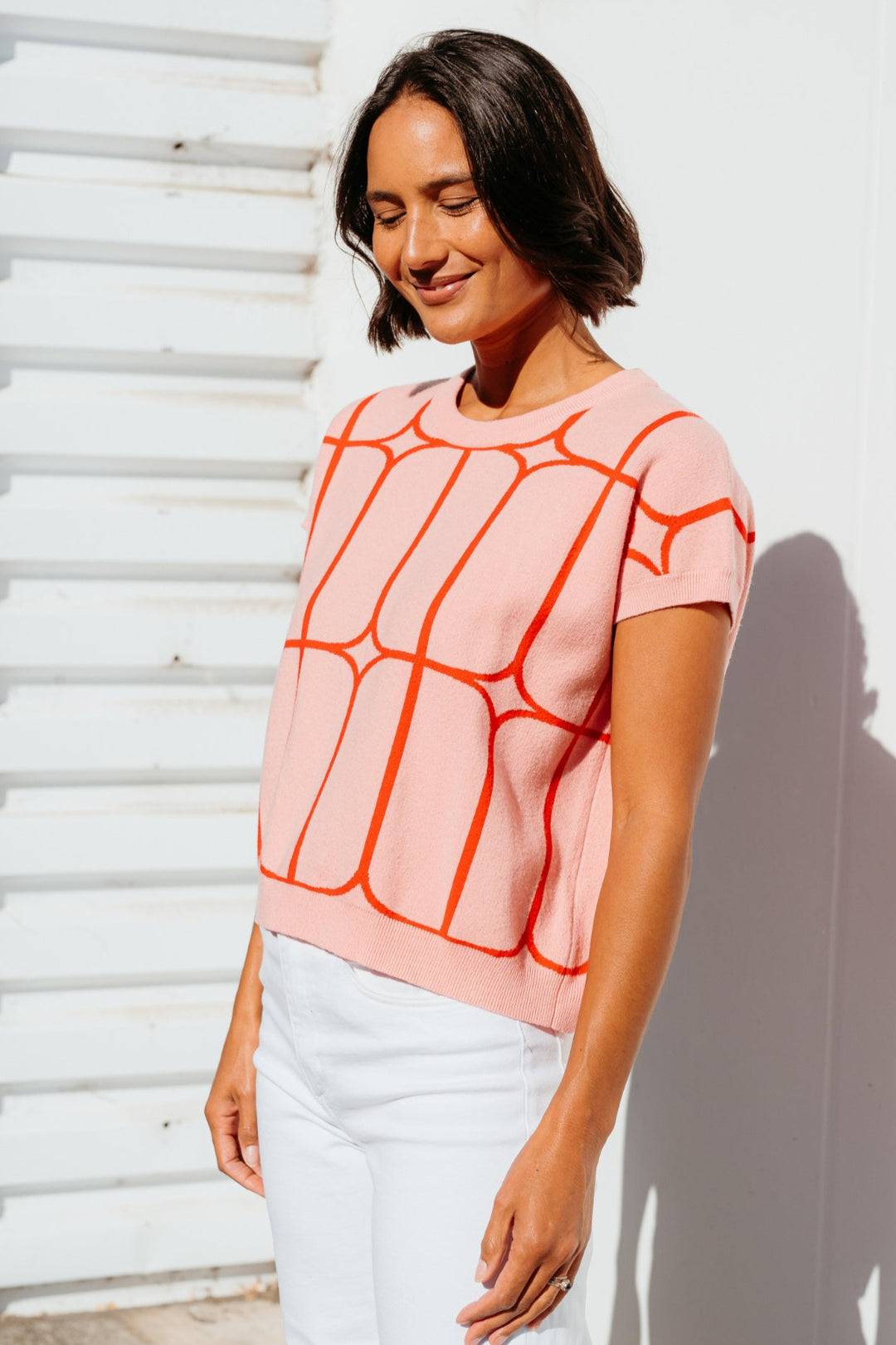 Woman wearing a pink top with orange geometric pattern standing against a white wall.