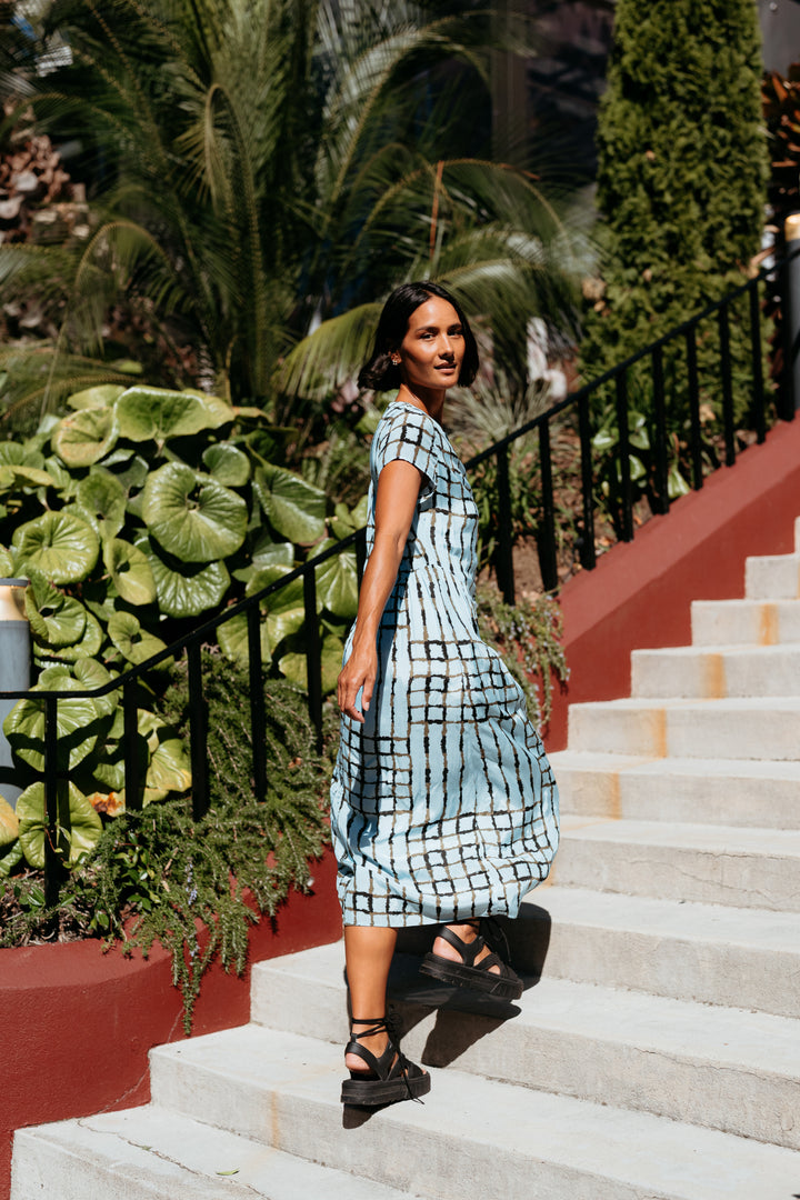 Woman in a checkered dress standing on stone steps with greenery in the background