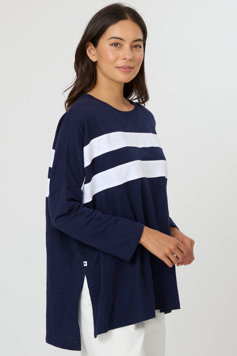 A woman modeling a navy blue long-sleeve top with two bold white horizontal stripes across the chest, paired with white pants, against a plain light gray studio background.