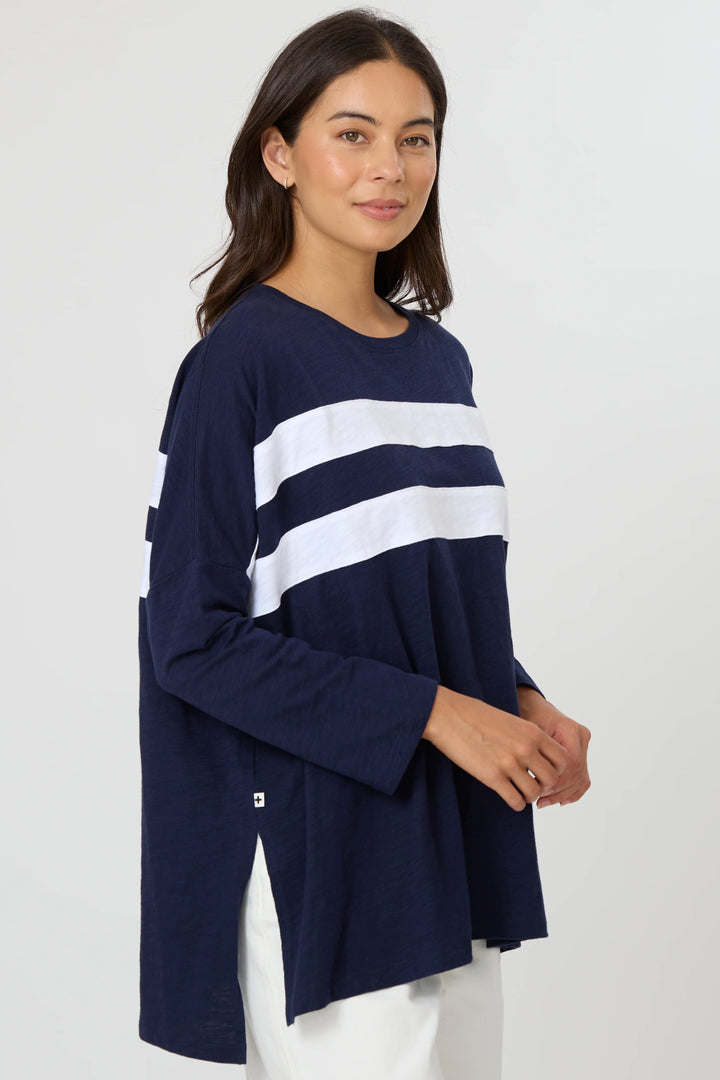 A woman modeling a navy blue long-sleeve top with two bold white horizontal stripes across the chest, paired with white pants, against a plain light gray studio background.