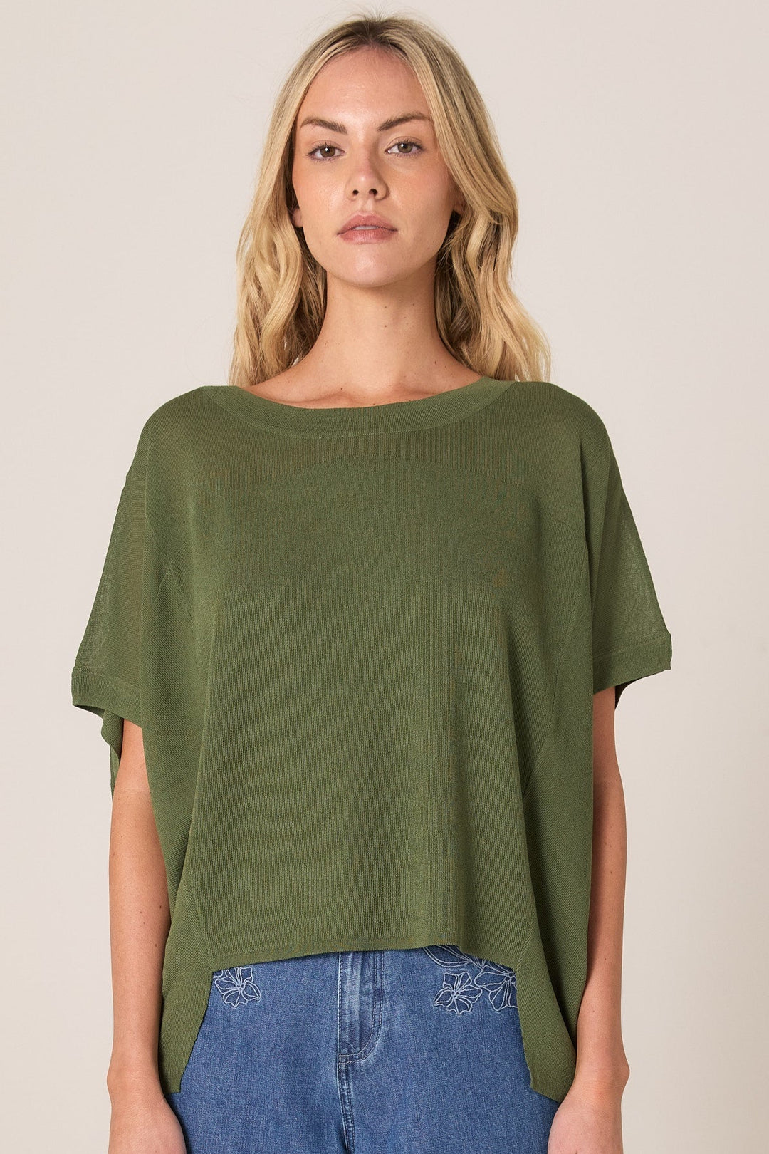 Woman wearing an olive green FATE & BECKER KNIT top and blue jeans on a beige background
