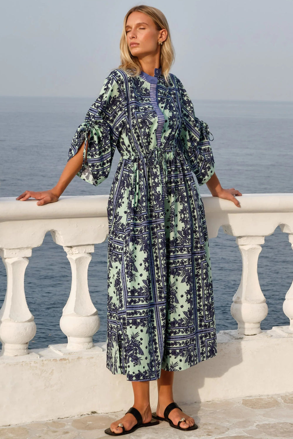 Woman in a patterned dress standing on a balcony with ocean view