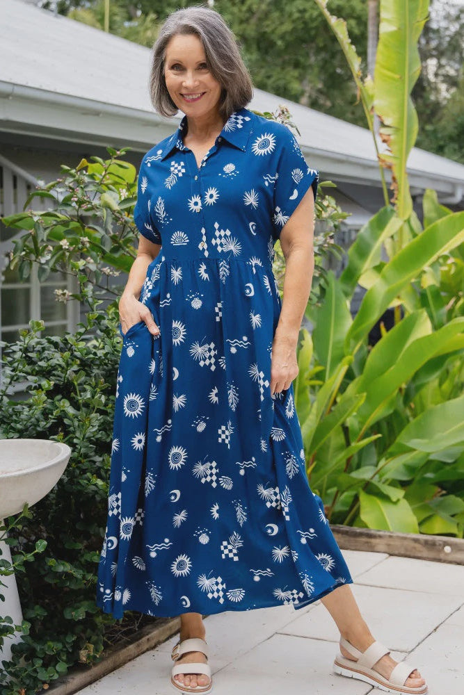 Woman wearing a blue floral dress standing outdoors with greenery around.