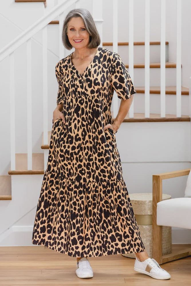 Woman wearing a leopard print dress standing in a home setting with stairs and a chair.