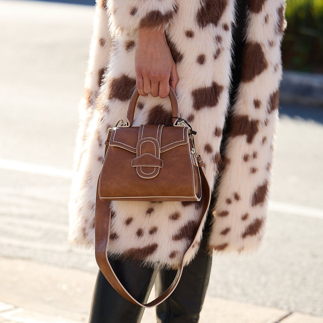Person wearing a long coat with a pattern, holding a brown handbag on a street.