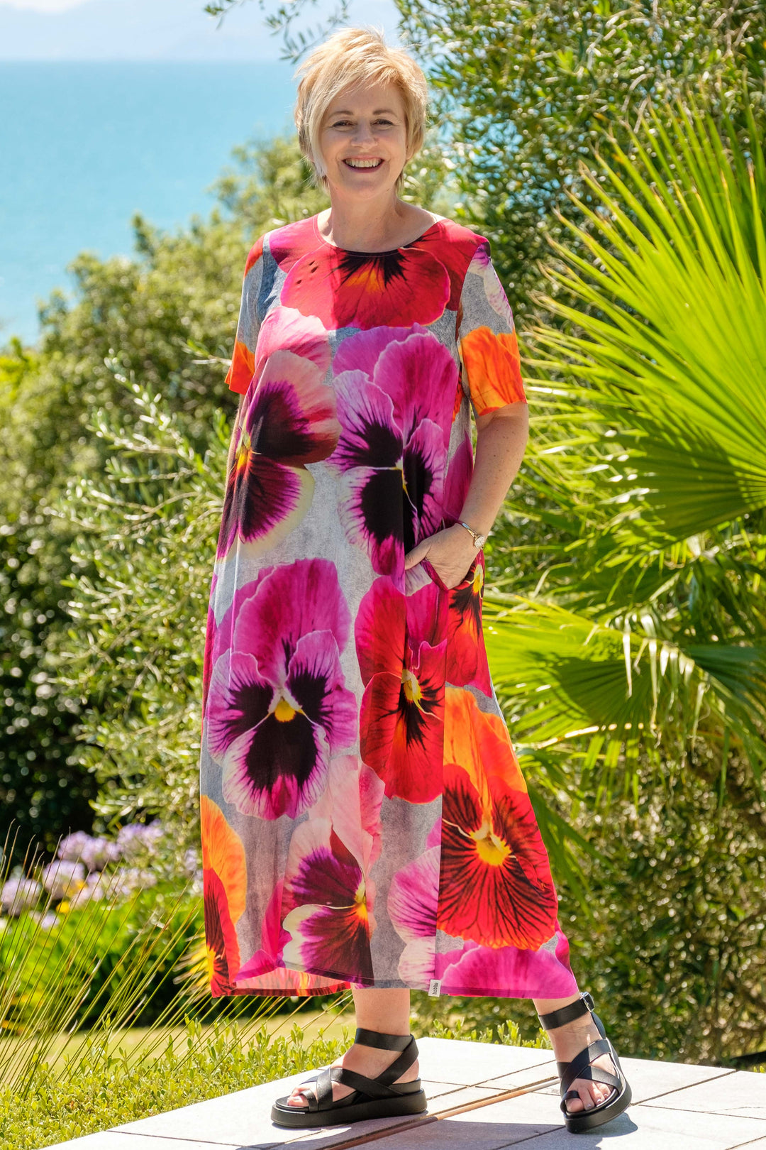 Woman in a colorful Deeanne Hobbs Menaka dress standing outdoors with greenery and blue sky in the background