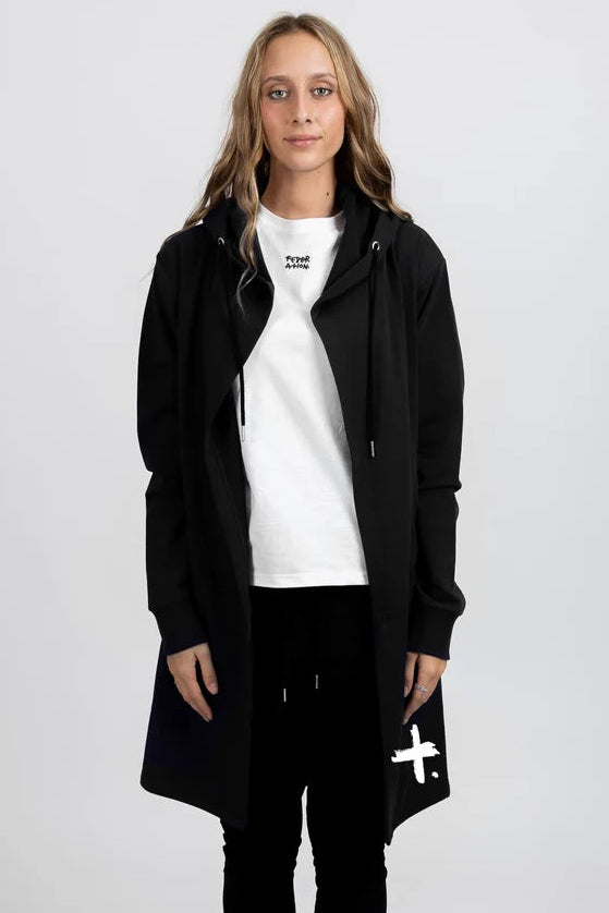 A model stands against a light gray studio background wearing a black hooded jacket over a white t-shirt, black joggers, and black-and-white sneakers. The jacket has a white cross graphic on the lower left hem and a small black logo on the chest.