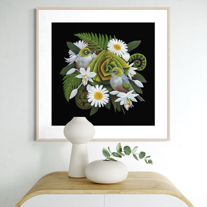 Framed artwork with green birds and flowers on a black background, displayed above a wooden cabinet.
