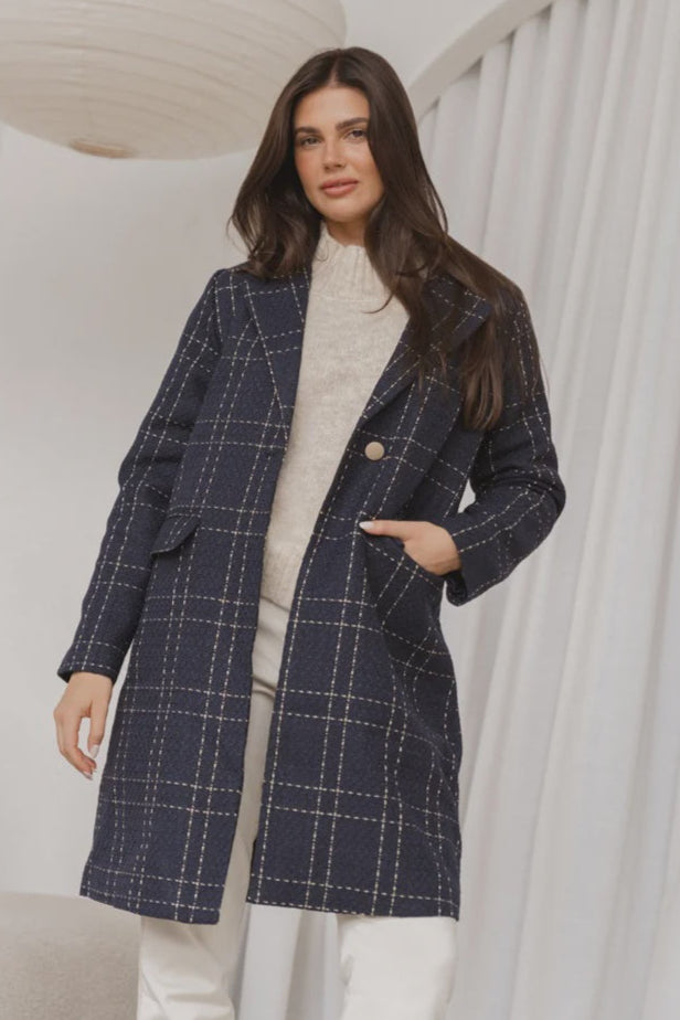 A female model wearing a long navy coat with a checkered pattern, featuring a classic collar and front flap pockets.