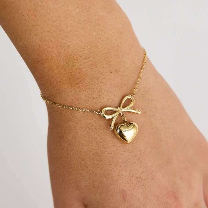 Gold bracelet with heart and bow charm on a wrist against a light background