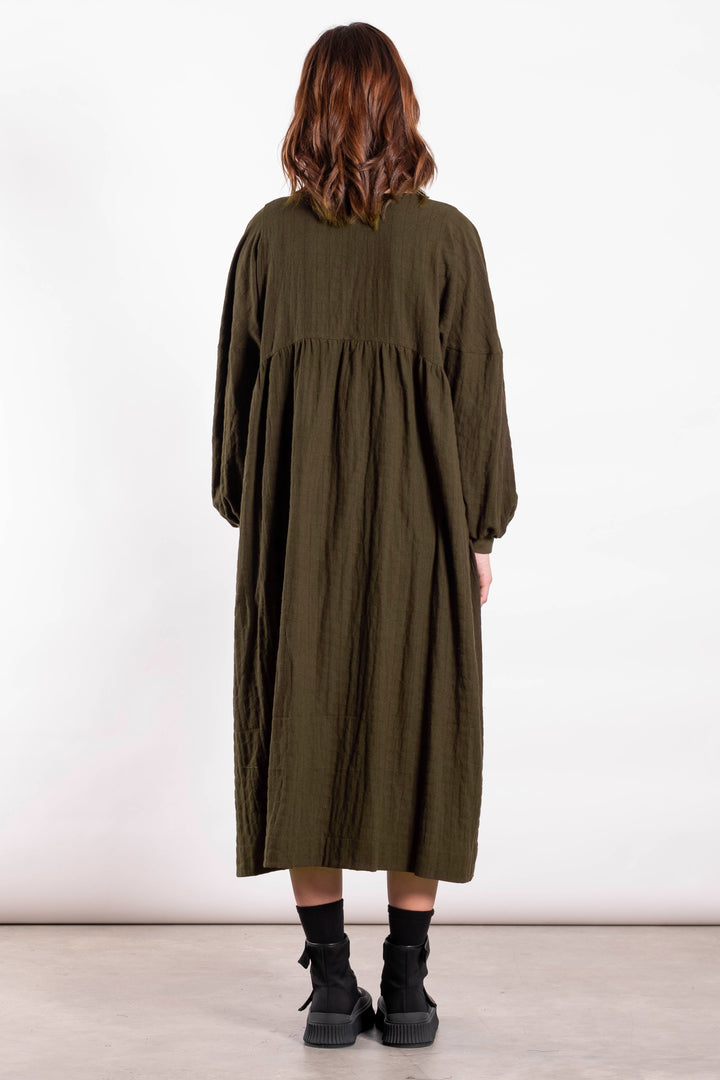 Person wearing a long green dress standing against a white wall.