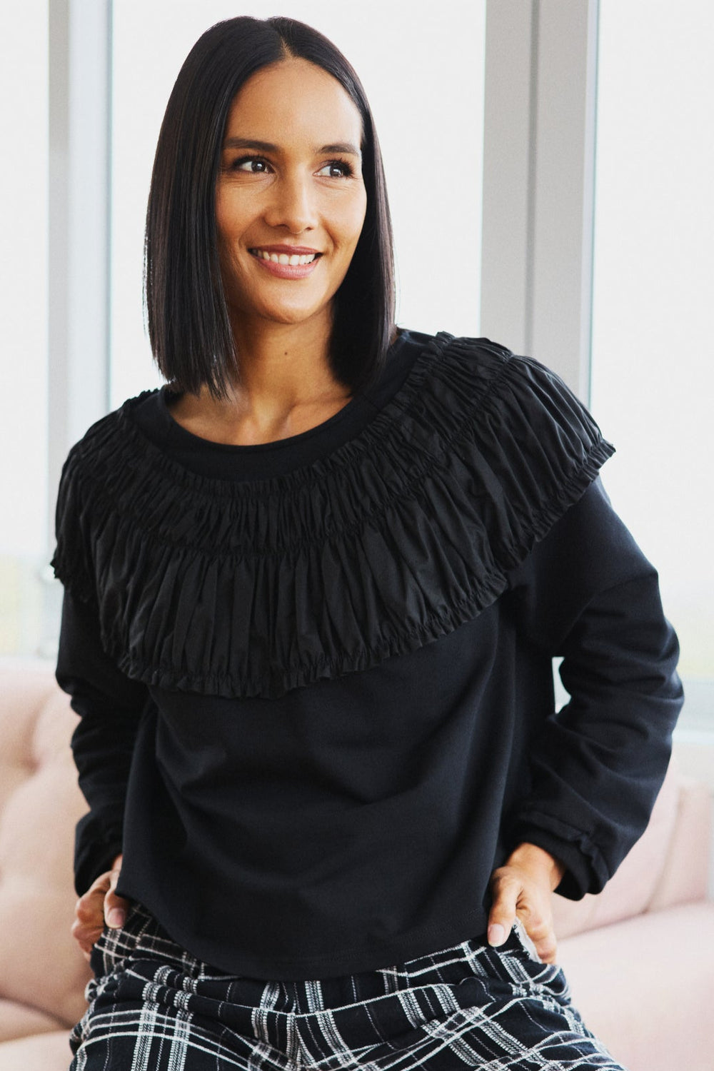 Woman wearing a black fringed sweater and plaid pants sitting on a pink couch.