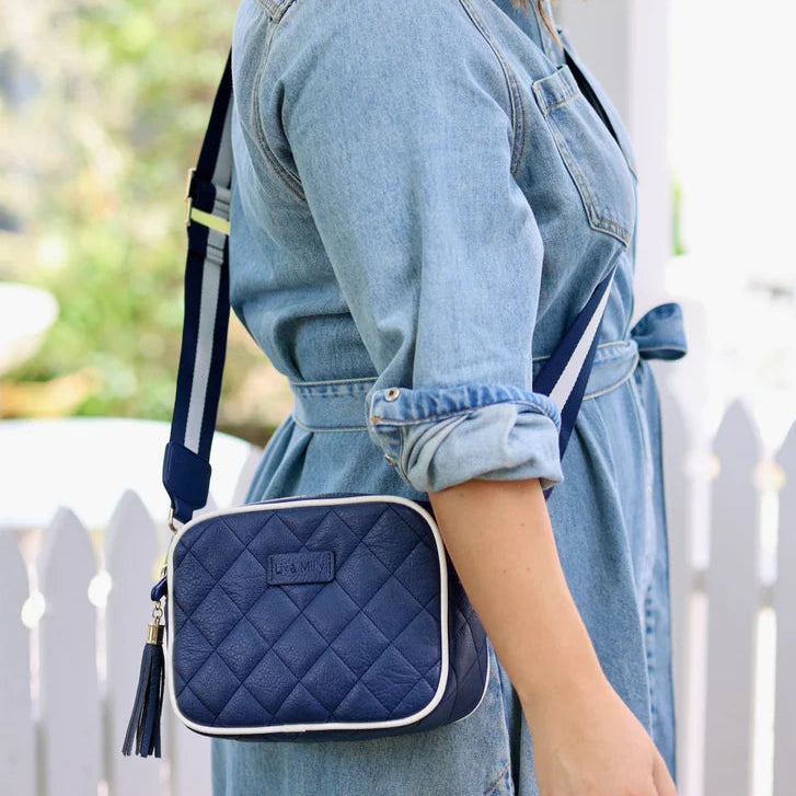 Woman wearing a denim dress with a navy quilted crossbody bag in front of a white picket fence.
