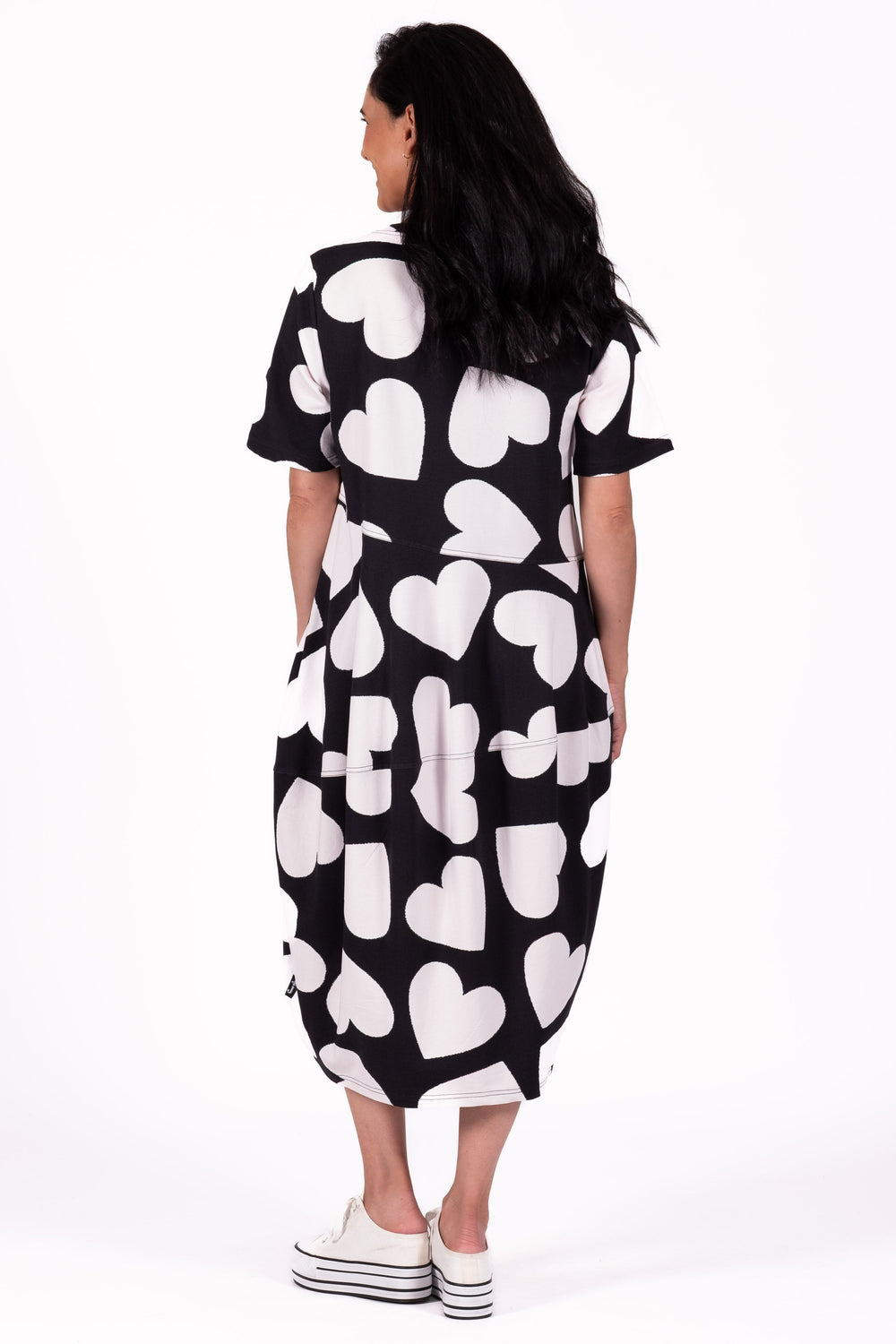 A woman viewed from behind wearing a short-sleeve midi dress with a black and white heart pattern, paired with white platform sneakers, against a plain white studio background.