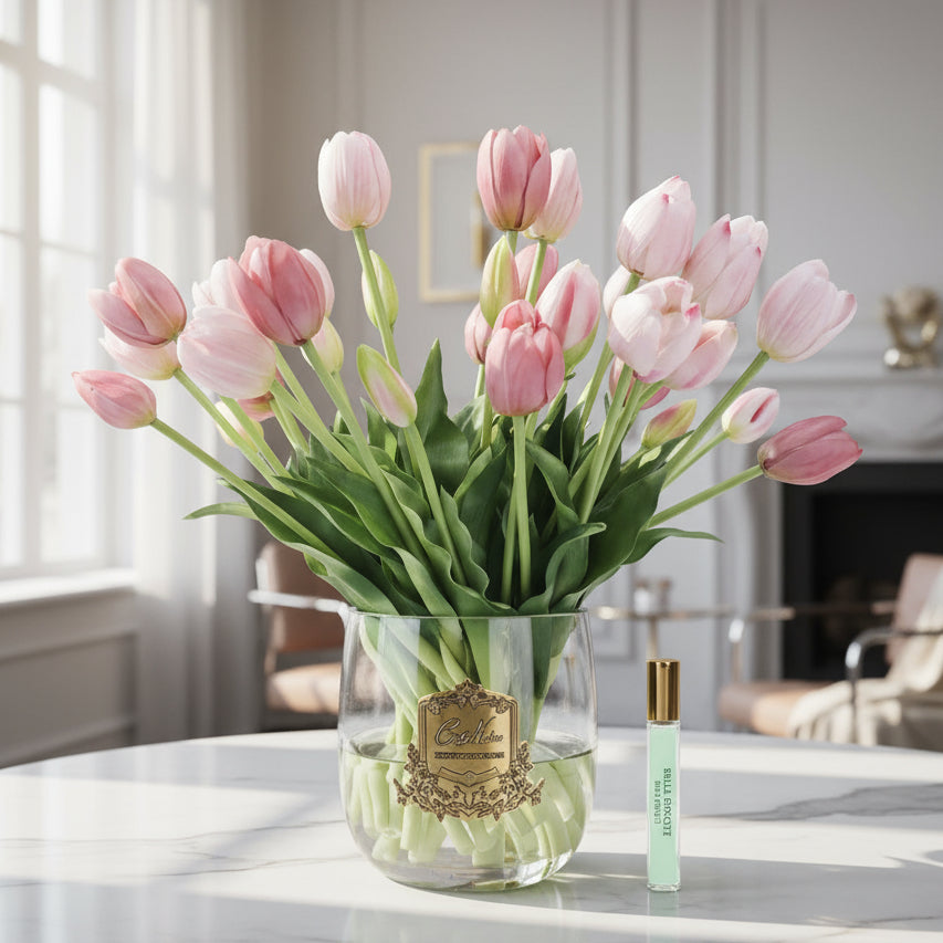 Bouquet of pink cote noire diffuser tulips in a clear vase with a small bottle on a white background