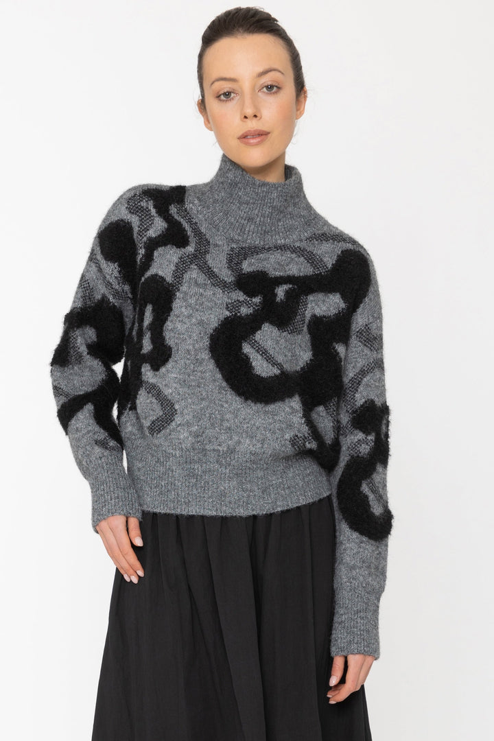 Person wearing a gray sweater with black abstract patterns and a black skirt on a white background