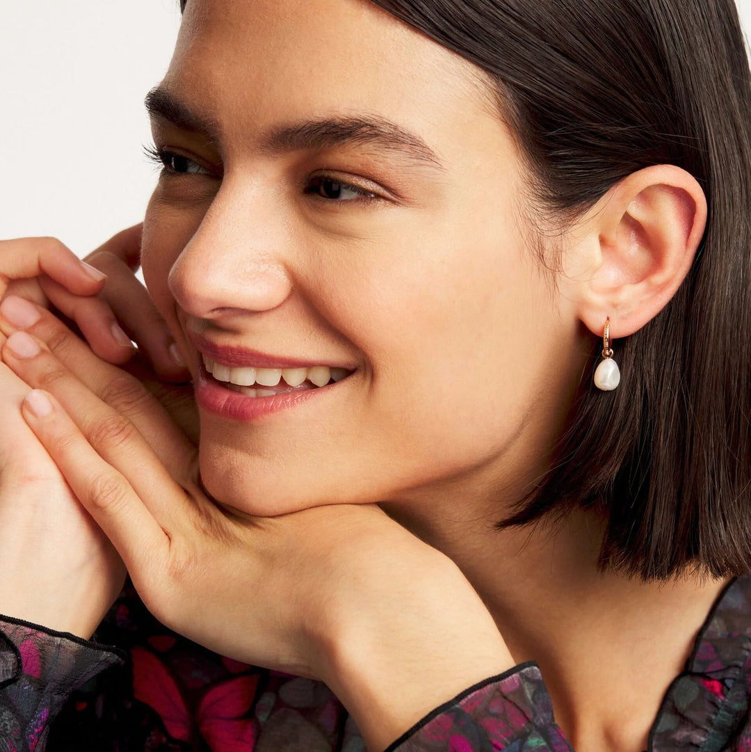 Woman with short dark hair wearing a floral blouse and ted baker pearl gold earrings, smiling with hands under chin.