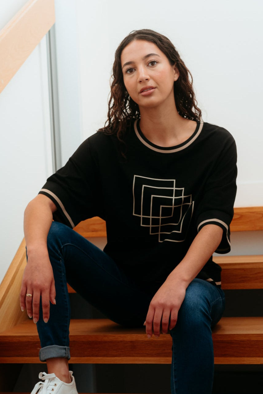 Person sitting on wooden stairs wearing a black sweater with a geometric design and white sneakers.