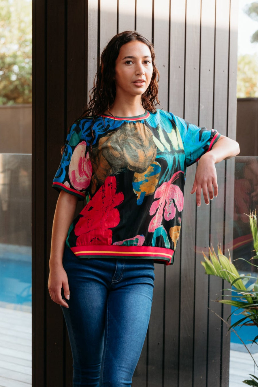 Woman wearing a colorful floral top and blue jeans standing on a wooden deck.