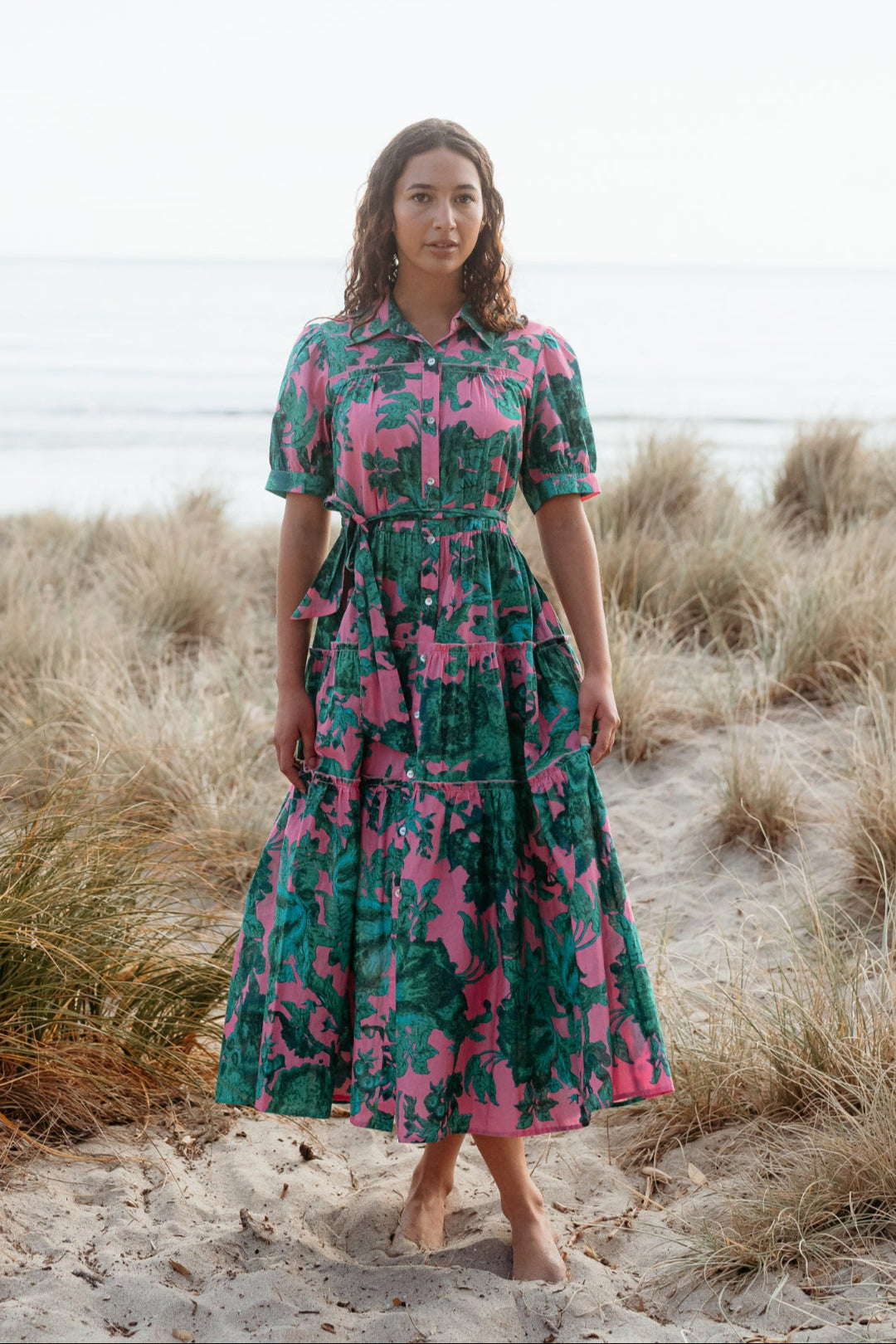 Woman in a patterned dress standing in a desert-like environment with dry grass and a white fence in the background.
