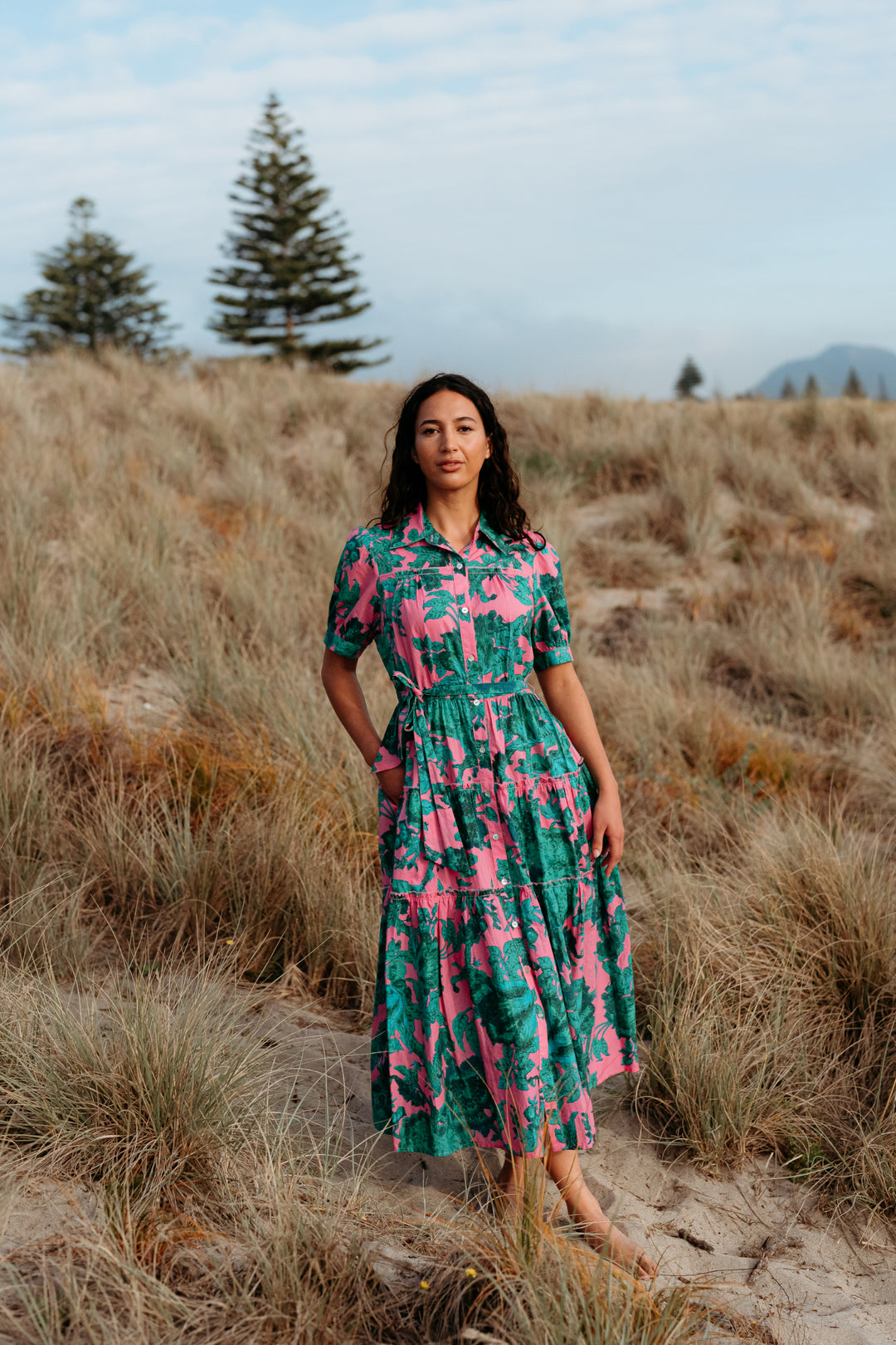 Woman in a patterned dress standing in a field with trees and mountains in the background