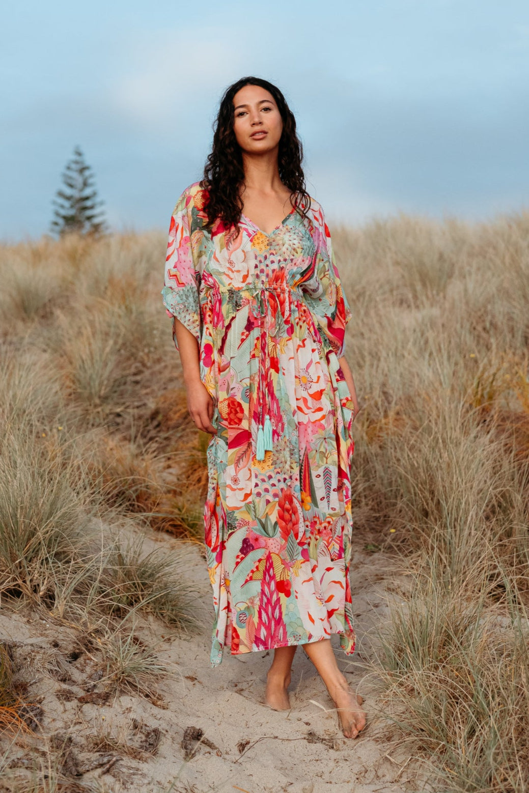 Woman in a floral dress standing in a sandy field with a blue sky.