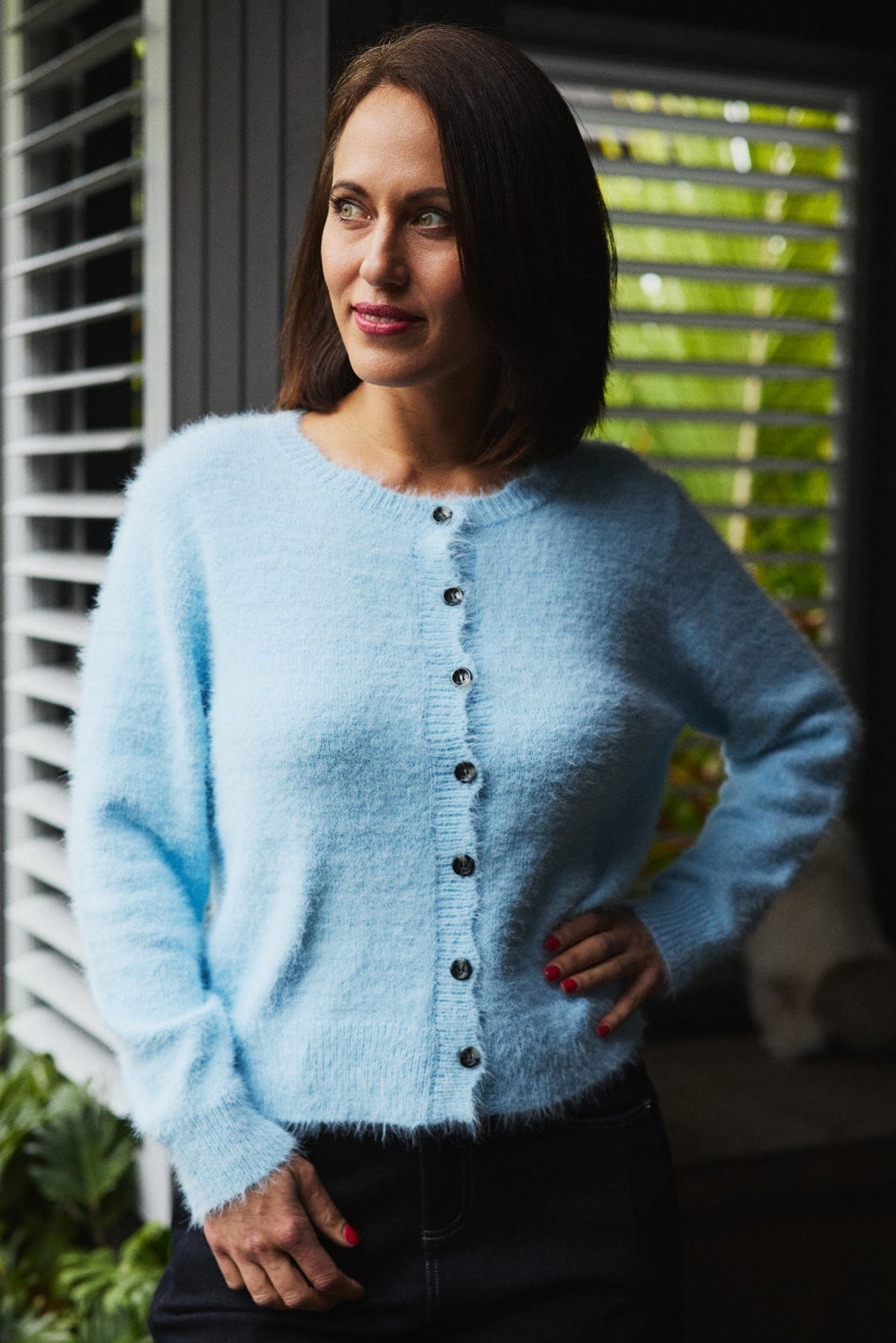 Woman wearing a light blue cardigan standing outdoors with greenery in the background