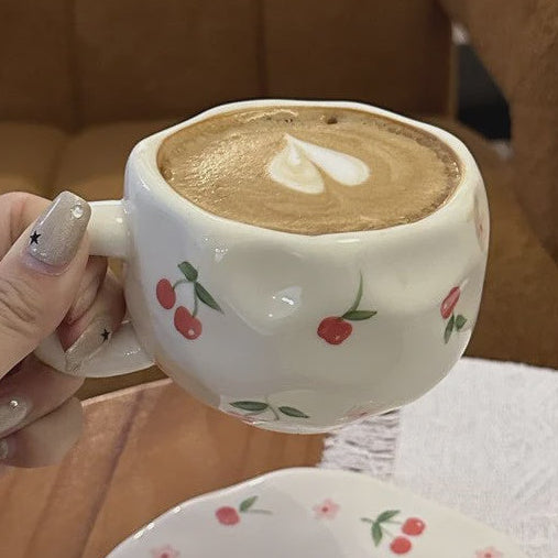 Person holding a ceramic coffee cup with cherry design on a table.