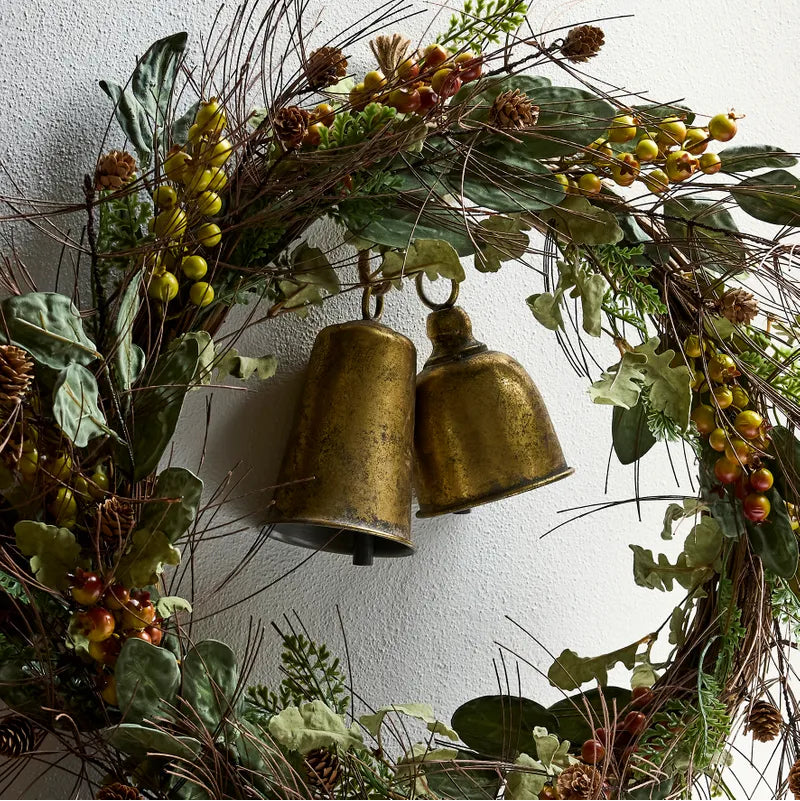 Decorative wreath with greenery, berries, and brass bells against a white wall.