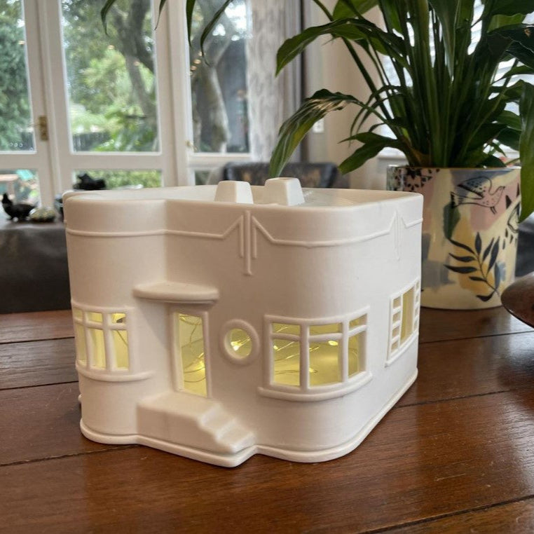 White ceramic house-shaped lantern on a wooden table with a plant in the background