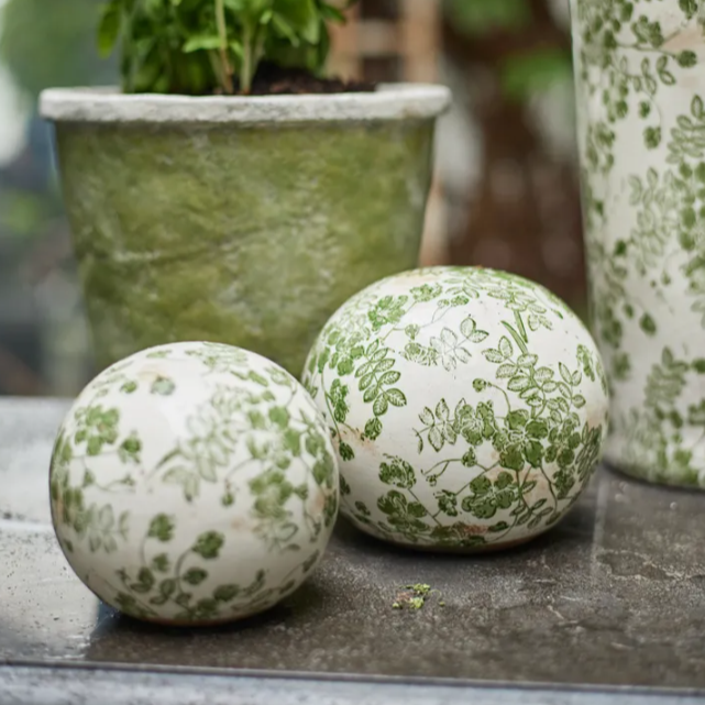 Two decorative balls with green floral patterns on a surface, with a blurred plant pot in the background.