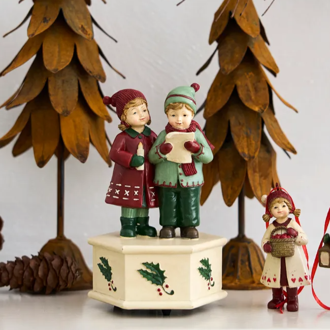 Decorative figurines of children with a music box on a white surface with wooden trees in the background.