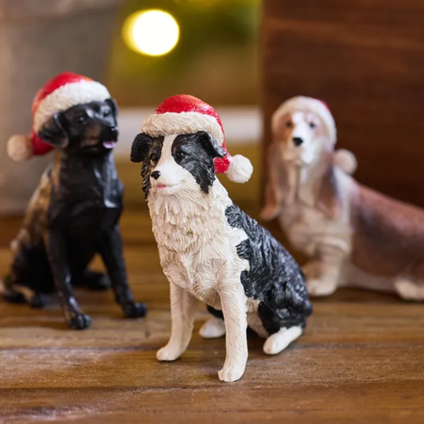 Three dog figurines wearing Santa hats on a wooden surface with a blurred Christmas tree in the background.