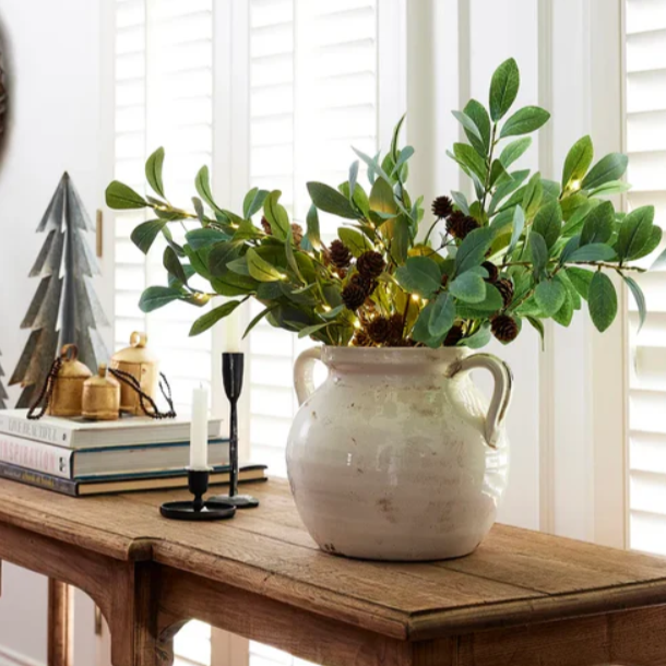 Decorative arrangement of greenery in a white vase on a wooden table with books and a small tree decoration.