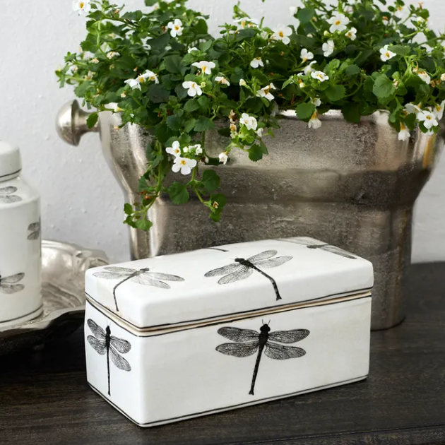 Decorative box with dragonfly design on a wooden surface with a metal planter of flowers in the background.