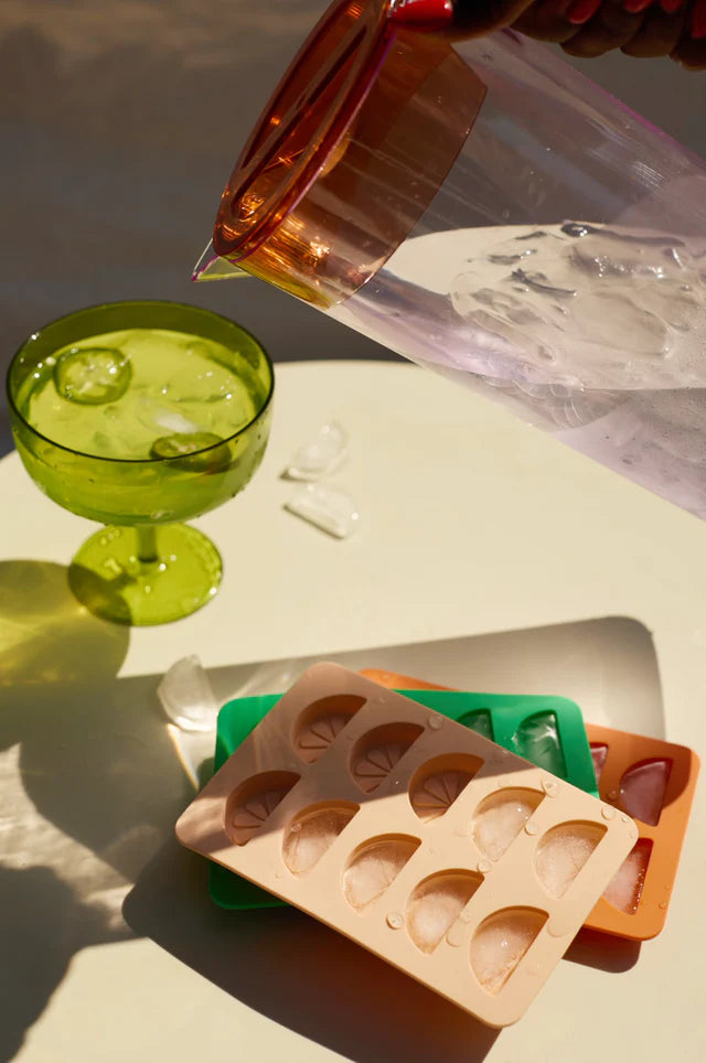 Person pouring a drink into a green glass with ice cubes, next to a silicone mold on a light surface.