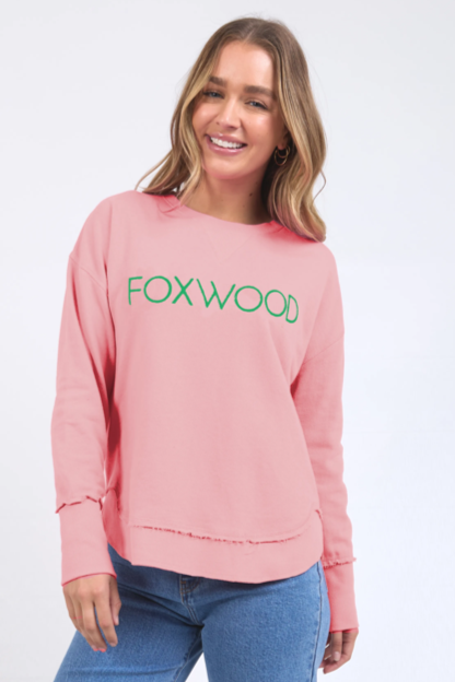 Woman wearing a pink sweatshirt with 'FOXWOOD' text, blue jeans, and white sneakers on a white background
