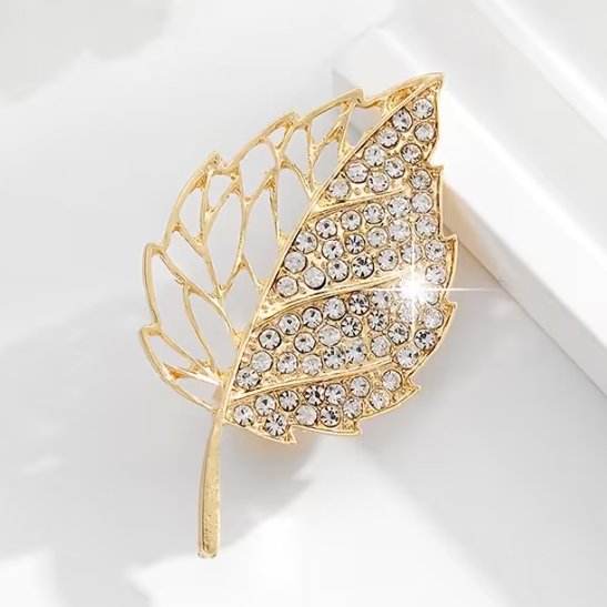 A gold-toned leaf-shaped brooch or pin embellished with clear rhinestones, photographed on a clean white surface with soft shadows.