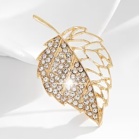A gold-toned leaf-shaped brooch embellished with clear rhinestones, displayed on a white fabric surface.