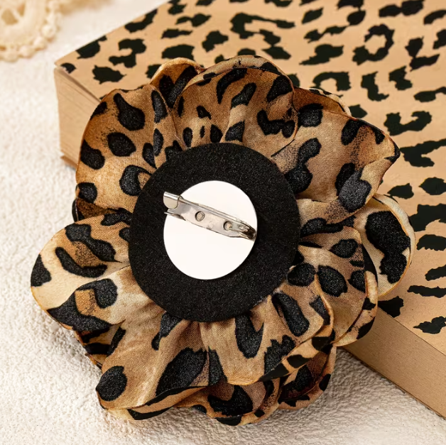 A leopard print fabric flower pin-back brooch with a black center and silver metal clasp, displayed on a textured white surface next to a leopard print box.