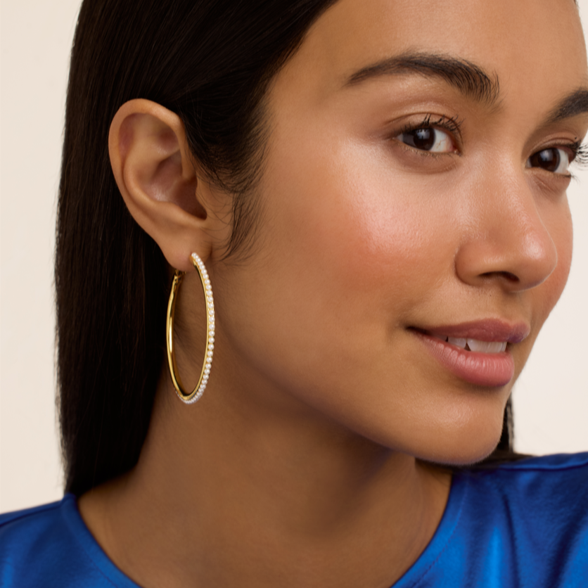 Woman wearing Ted Baker gold hoop earrings with a blue top against a beige background