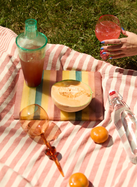 Outdoor setting with a halved cantaloupe on a striped blanket, drinks, coktail shaker, and a hand holding a glass.