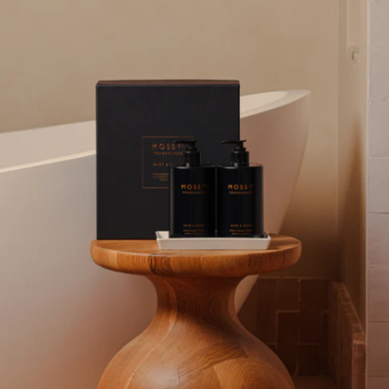 Wooden side table with black MOSS ST HAND CARE bottles and a box in front of a bathtub.