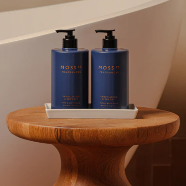 Two blue MOSS ST HAND CARE bottles with black pumps on a wooden table in front of a bathtub.