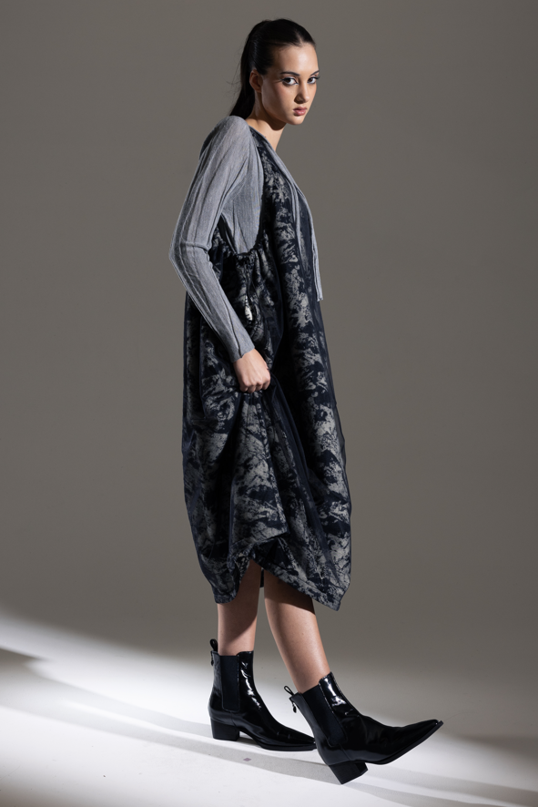 Woman wearing a long, dark patterned dress with a gray cardigan against a neutral background