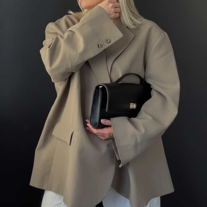 A woman stands indoors against a black paneled wall and white window trim, wearing a light beige oversized blazer, loose white pants, and holding a small black handbag.