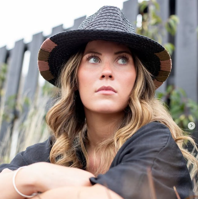 A woman wearing a black woven wide-brim hat with brown and tan striped side panels, looking upward outdoors against a dark fence and plants.